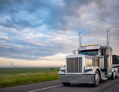 Gray classic big rig American semi truck with turned on lights and chrome exhaust pipes transporting covered cargo on flat bed semi trailer driving on the twilight highway road with stormy clouds sky